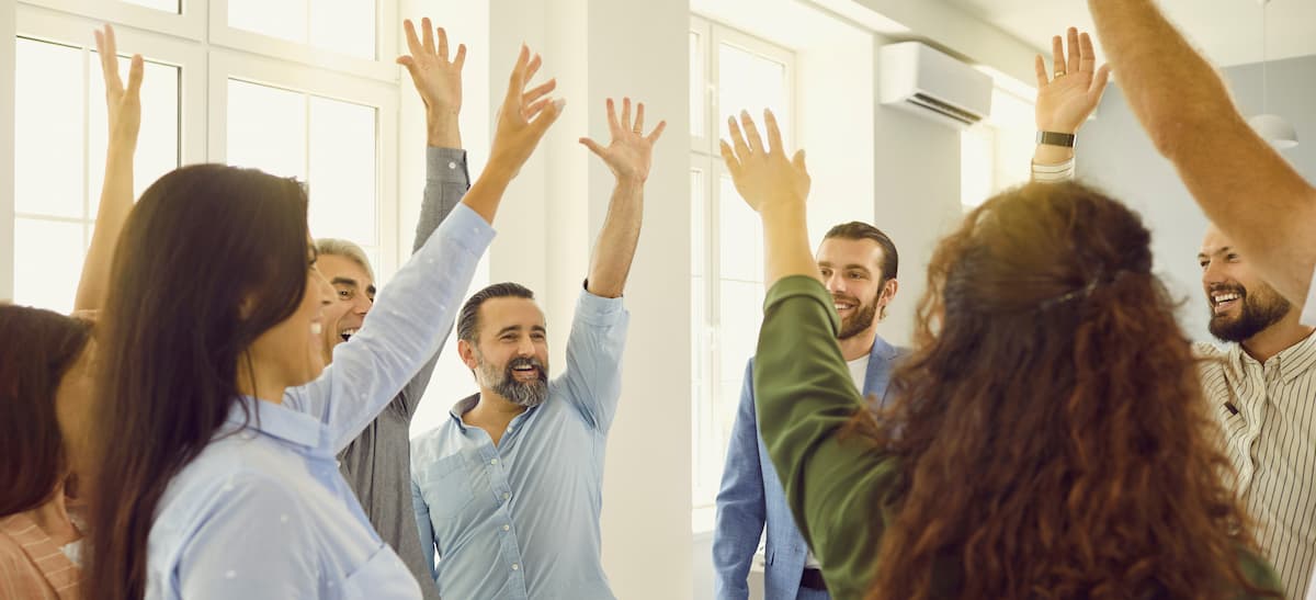 group of excited, smiling people with their arms up in the air standing in a circle inside a workshop room